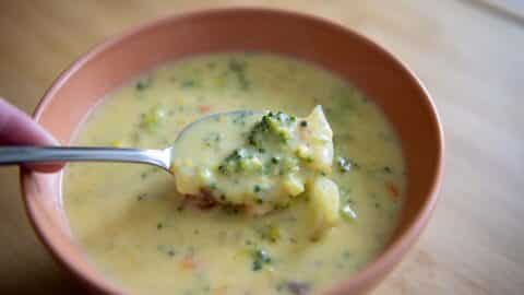 A spoonful of creamy broccoli cheddar soup is held above a bowl, showing chunks of broccoli and carrots in a thick yellow broth—an irresistible Broccoli Cheddar Soup Recipe come to life.