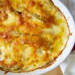 Golden-brown, bubbly cheese topping on Delicious Homemade Au Gratin Potatoes in a white oval baking dish, with a white cloth beside it on a wooden surface.
