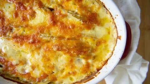 Golden-brown, bubbly cheese topping on Delicious Homemade Au Gratin Potatoes in a white oval baking dish, with a white cloth beside it on a wooden surface.