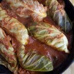 A black skillet filled with Töltött Káposzta (Cabbage Rolls) simmering in tomato sauce rests on a light wooden surface. The cabbage leaves are soft, covered with savory pieces of tomato and sauce.