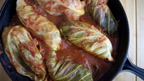 A black skillet filled with Töltött Káposzta (Cabbage Rolls) simmering in tomato sauce rests on a light wooden surface. The cabbage leaves are soft, covered with savory pieces of tomato and sauce.