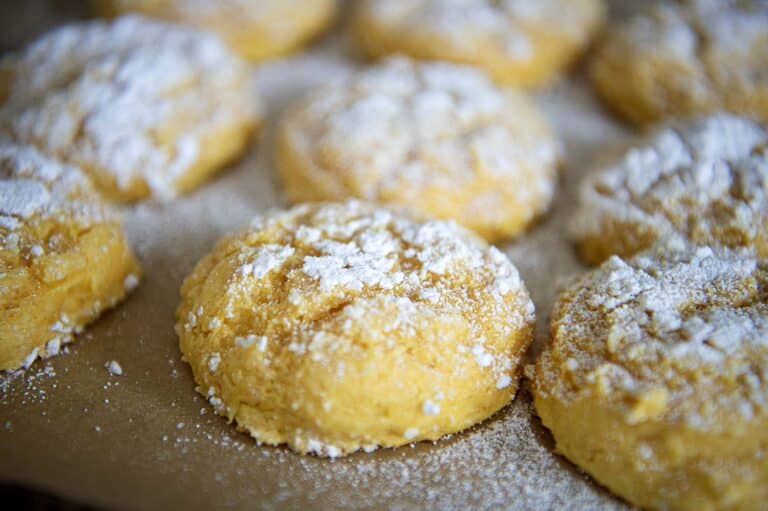 Close-up of several round, golden Pumpkin Gooey Butter Cookies dusted with powdered sugar, arranged on a sheet of parchment paper. The cookies have a soft, textured surface and are closely grouped together.