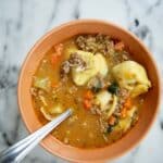A bowl of Sausage Tortellini Soup with ground meat, carrots, and herbs, with a spoon resting inside the bowl, placed on a white marble surface.