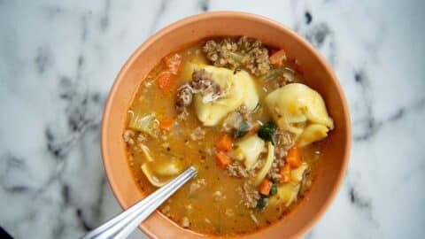 A bowl of Sausage Tortellini Soup with ground meat, carrots, and herbs, with a spoon resting inside the bowl, placed on a white marble surface.