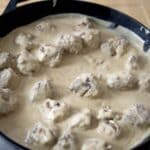 A cast iron skillet filled with köttbullar—Swedish meatballs—covered in a creamy white sauce, resting on a light wooden surface.