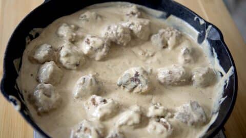 A cast iron skillet filled with köttbullar—Swedish meatballs—covered in a creamy white sauce, resting on a light wooden surface.