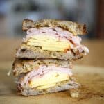 A stacked Vermonter Sandwich breakfast with scrambled eggs and sliced turkey or ham between two pieces of toasted rustic bread, sitting on a wooden cutting board.