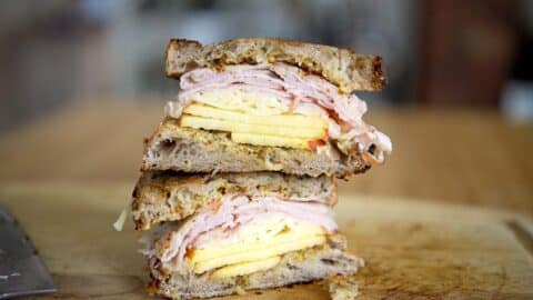 A stacked Vermonter Sandwich breakfast with scrambled eggs and sliced turkey or ham between two pieces of toasted rustic bread, sitting on a wooden cutting board.