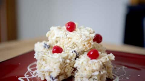 Four white, crispy dessert squares from a classic White Christmas Recipe are stacked on a red plate, topped with bright red cherries and drizzled with white icing. The softly blurred background highlights their festive appeal.