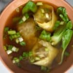 A bowl of Wonton Soup with green onions and bok choy sits in a clear broth, shown from above on a white surface.