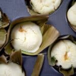 Close-up of Bibingka rice cakes with cracked tops, each wrapped in banana leaves and placed in a metal muffin tin. The cakes appear light, fluffy, and freshly baked.