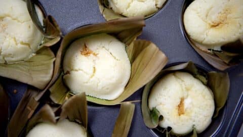 Close-up of Bibingka rice cakes with cracked tops, each wrapped in banana leaves and placed in a metal muffin tin. The cakes appear light, fluffy, and freshly baked.