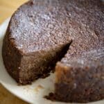A round, dense Jamaican Black Cake with a single slice cut out sits on a white plate atop a wooden surface. The cake appears moist with a slightly glossy, textured surface.