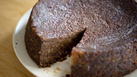 A round, dense Jamaican Black Cake with a single slice cut out sits on a white plate atop a wooden surface. The cake appears moist with a slightly glossy, textured surface.