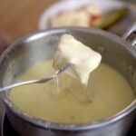 A fork holds a piece of bread dipped in melted Swiss Fondue cheese above a pot, with a blurred plate of food in the background.