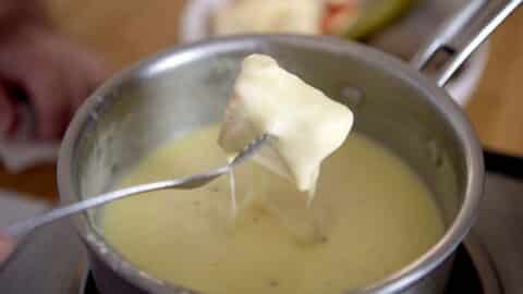 A fork holds a piece of bread dipped in melted Swiss Fondue cheese above a pot, with a blurred plate of food in the background.