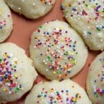 Close-up of round Lemon Ricotta Cookies topped with colorful rainbow sprinkles, arranged on an orange surface. The cookies have a glossy glaze and are closely placed together.
