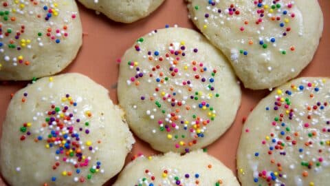 Close-up of round Lemon Ricotta Cookies topped with colorful rainbow sprinkles, arranged on an orange surface. The cookies have a glossy glaze and are closely placed together.