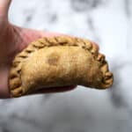 A hand holding a baked Nevri empanada with a braided edge, set against a blurred white and gray background.