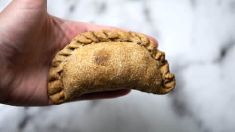 A hand holding a baked Nevri empanada with a braided edge, set against a blurred white and gray background.
