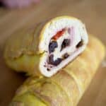 A close-up of a sliced Pan de Jamon Venezolano displays its golden-brown crust and a savory filling of ham, raisins, and olives on a wooden surface.