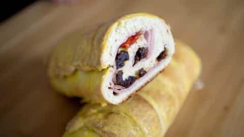 A close-up of a sliced Pan de Jamon Venezolano displays its golden-brown crust and a savory filling of ham, raisins, and olives on a wooden surface.