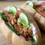 Two sandwich rolls filled with shredded seasoned meat, lettuce, sliced cucumber, tomato, and radish, placed on a wooden surface.