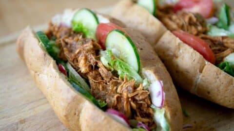 Two sandwich rolls filled with shredded seasoned meat, lettuce, sliced cucumber, tomato, and radish, placed on a wooden surface.