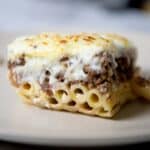 A close-up of a slice of pastitsio casserole with layers of tubular pasta, seasoned ground meat, and a thick, golden-brown layer of melted cheese on top, served on a beige plate.