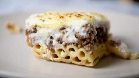 A close-up of a slice of pastitsio casserole with layers of tubular pasta, seasoned ground meat, and a thick, golden-brown layer of melted cheese on top, served on a beige plate.