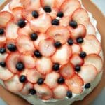 A pavlova dessert topped with overlapping slices of fresh strawberries and scattered blueberries sits on a light brown plate, seen from above on a marble surface—an irresistible pavlova recipe brought to life.