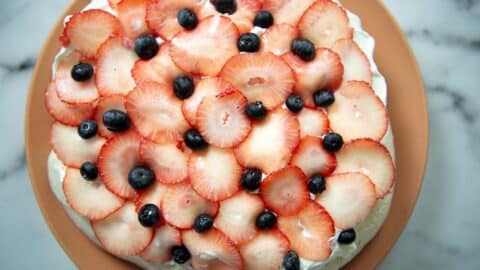 A pavlova dessert topped with overlapping slices of fresh strawberries and scattered blueberries sits on a light brown plate, seen from above on a marble surface—an irresistible pavlova recipe brought to life.