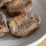 Three pieces of Rabanada coated in cinnamon sugar are arranged on a white plate, with a golden-brown, crispy appearance. The classic Brazilian treat is beautifully presented on a light wooden surface.