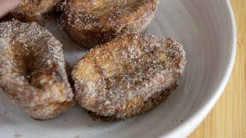 Three pieces of Rabanada coated in cinnamon sugar are arranged on a white plate, with a golden-brown, crispy appearance. The classic Brazilian treat is beautifully presented on a light wooden surface.