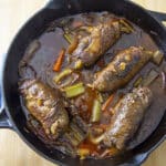 A cast iron skillet filled with tender Rouladen simmering in a rich brown sauce, surrounded by pieces of celery, carrots, and onions, rests on a light wooden surface.