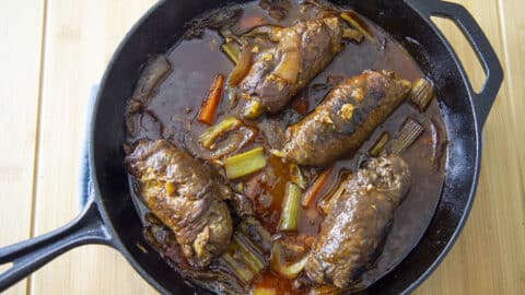 A cast iron skillet filled with tender Rouladen simmering in a rich brown sauce, surrounded by pieces of celery, carrots, and onions, rests on a light wooden surface.