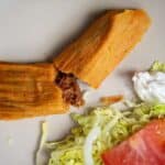 A plate featuring the Best Pork Tamales Rojos split in half to reveal savory shredded meat inside, served with shredded lettuce, a tomato slice, onion, and a dollop of sour cream.