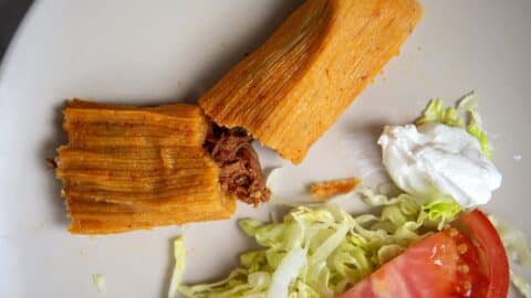 A plate featuring the Best Pork Tamales Rojos split in half to reveal savory shredded meat inside, served with shredded lettuce, a tomato slice, onion, and a dollop of sour cream.
