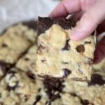 A hand holding a square chocolate chip cookie bar with a marbled chocolate and vanilla pattern; more brookie recipe bars are visible in the background.