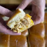 A person holds a Chicken Bacon Ranch Slider with pulled chicken and cheese on a soft, golden bun, above a tray of similar sliders.