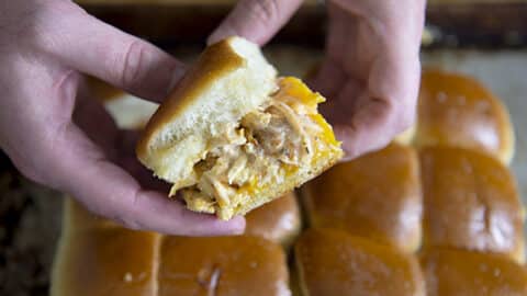 A person holds a Chicken Bacon Ranch Slider with pulled chicken and cheese on a soft, golden bun, above a tray of similar sliders.