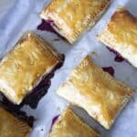 Six golden-brown rectangular hand pies, reminiscent of Cherry Toaster Strudel, are arranged on a sheet of parchment paper, with vibrant berry filling oozing from the sides.