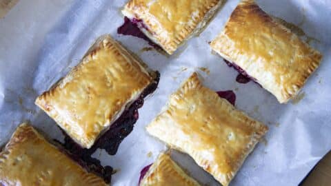 Six golden-brown rectangular hand pies, reminiscent of Cherry Toaster Strudel, are arranged on a sheet of parchment paper, with vibrant berry filling oozing from the sides.