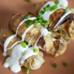 Three crispy, golden-brown Jalapeno Popper Egg Rolls topped with a drizzle of white sauce and sprinkled with chopped green onions, served on a peach-colored plate.