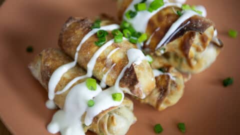 Three crispy, golden-brown Jalapeno Popper Egg Rolls topped with a drizzle of white sauce and sprinkled with chopped green onions, served on a peach-colored plate.