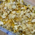 A casserole dish filled with The Best Old Fashioned Tuna Noodle Casserole, topped with crumbled potato chips, melted cheese, and visible pieces of vegetables, resting on a wooden surface.