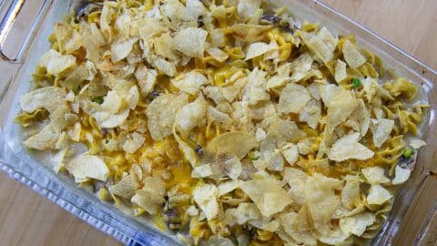 A casserole dish filled with The Best Old Fashioned Tuna Noodle Casserole, topped with crumbled potato chips, melted cheese, and visible pieces of vegetables, resting on a wooden surface.