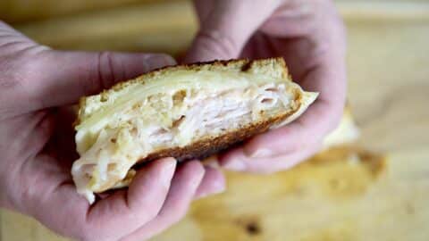 Close-up of hands holding a sliced Turkey Reuben sandwich with toasted bread, melted cheese, tender turkey, and sauerkraut on a wooden surface.