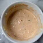 A white bowl filled with a creamy, light orange Turkey Reuben sauce speckled with small bits of seasoning sits on a marble surface. Some plastic wrap is partially visible next to the bowl.