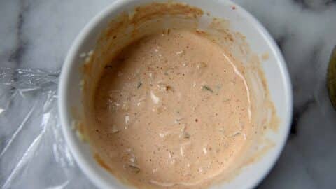 A white bowl filled with a creamy, light orange Turkey Reuben sauce speckled with small bits of seasoning sits on a marble surface. Some plastic wrap is partially visible next to the bowl.